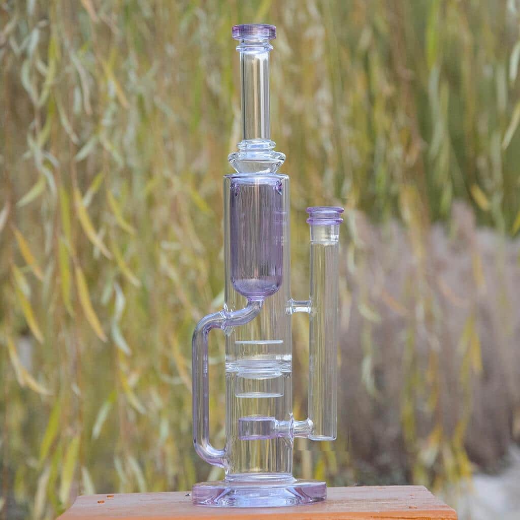 Front view of the FLOWER KLEIN BONG, a pale purple glass piece with a streamlined design, against a blurred green background.