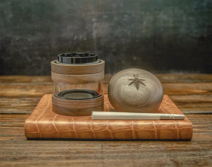 A top-down shot shows The Bzz 4 Piece Grinder resting on a textured mat, with its metallic grinding teeth partially visible.