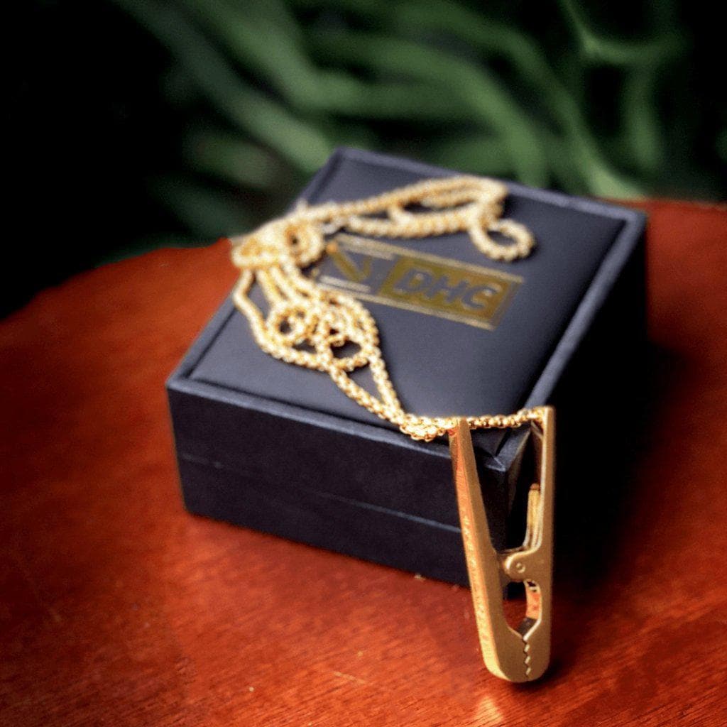 A close-up shot of the gold Daily High Club Roach Clip resting on its branded black box, showcasing its sleek, metallic design.