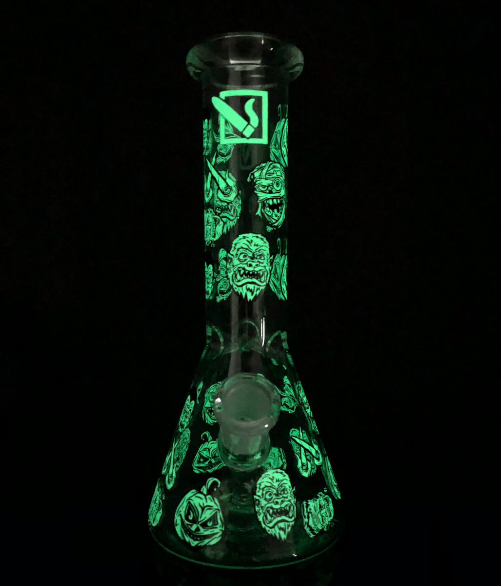 Front view of the DHC Originals "Monster Mash Beaker" Bong, featuring monster faces illustrated on the clear glass.