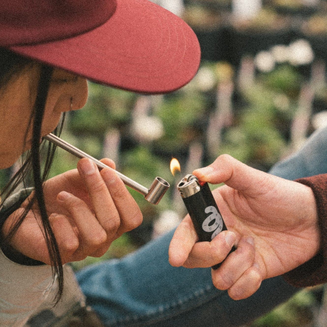 A close-up shows a hand lighting the bowl of the Long Cobb Titanium Pipe, with a blurred outdoor background.