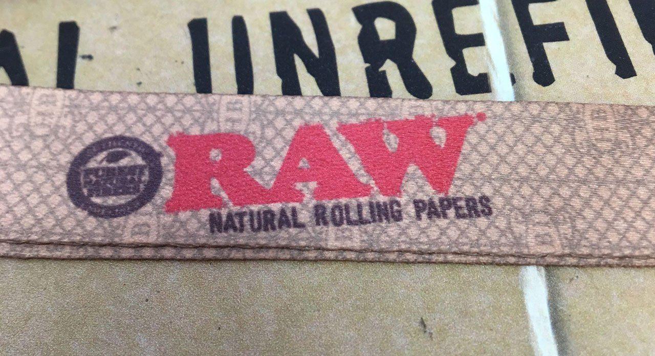 Close-up of the Raw Lanyard + Roach Clip featuring bold lettering against a textured, dark background.