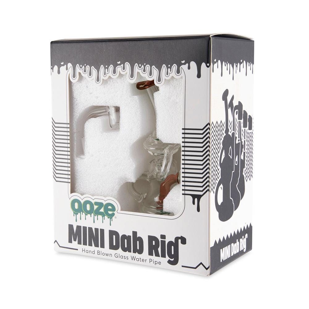A side view of the Ooze Surge Mini Recycler Dab Rig, showcasing its clear glass construction against a dark, patterned backdrop.