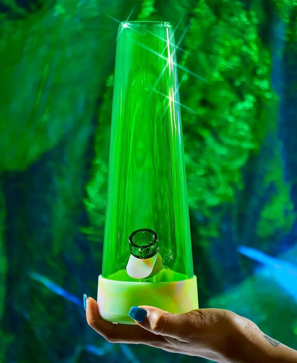 A hand holds the Designer Bong GLOW, illuminated with a vibrant, glowing effect against a blurred green backdrop.