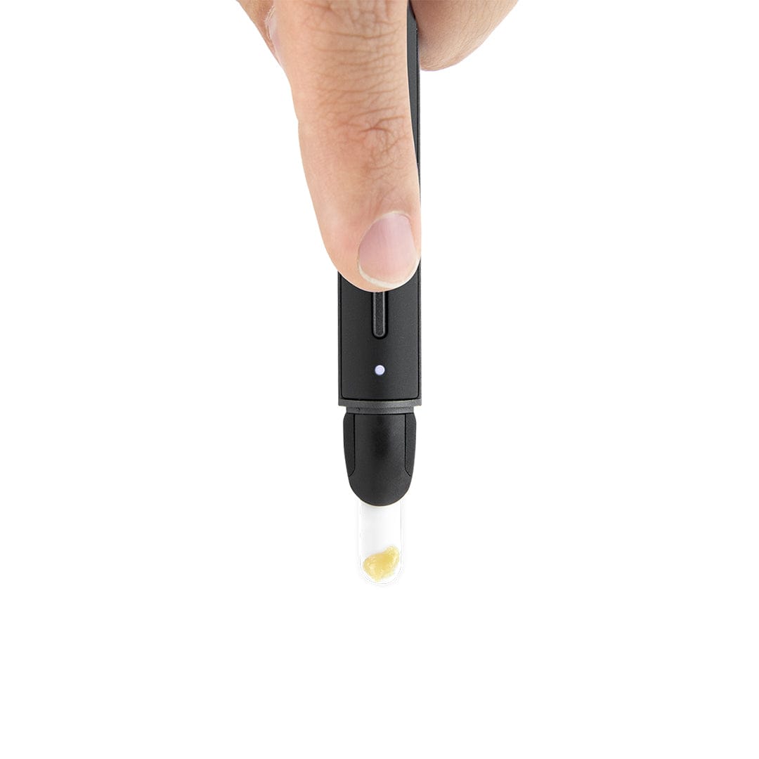 Top-down close-up of a finger pressing the black Puffco New Hot Knife, showing its LED indicator and a small glass tip with a dab.