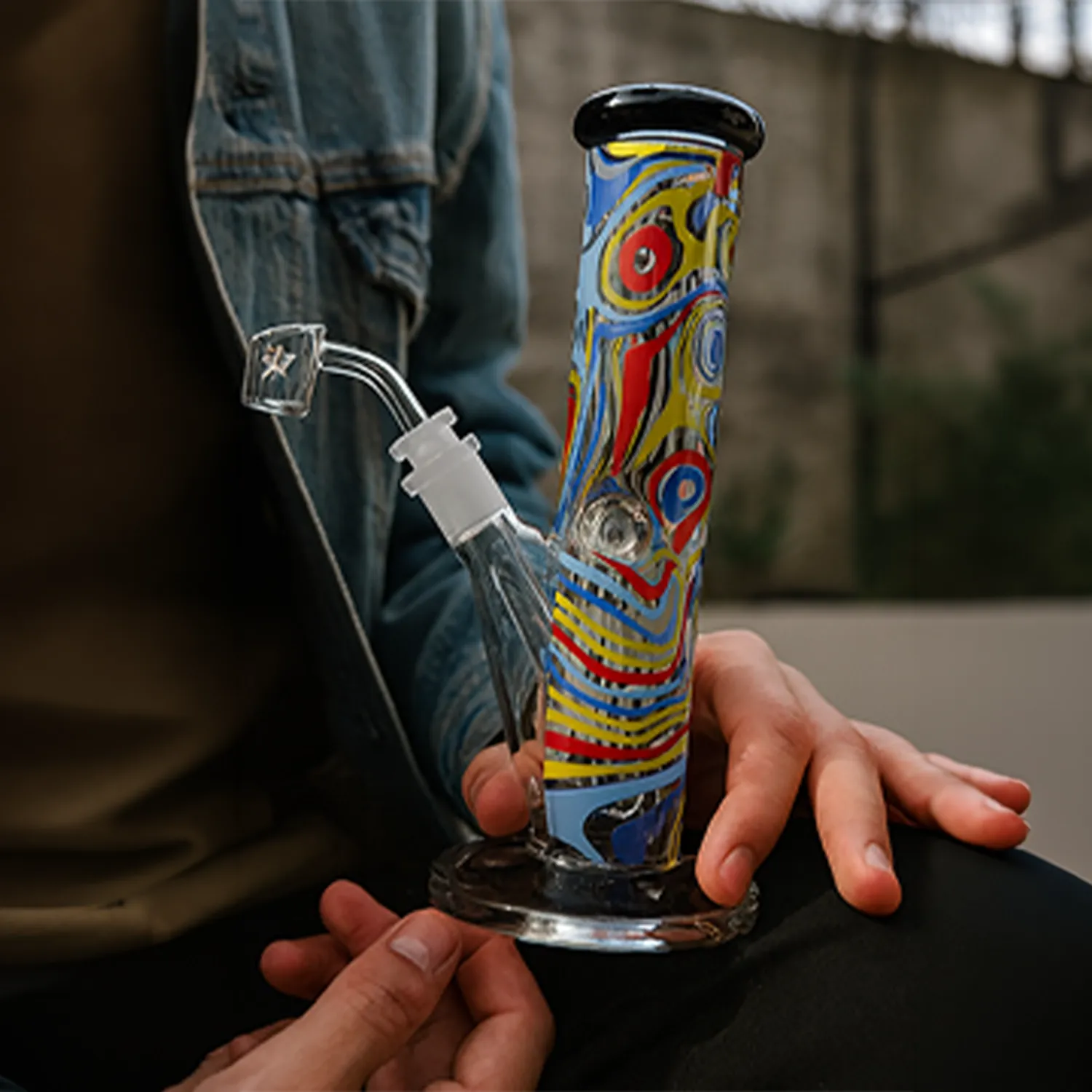 Close-up of a person holding the colorful, swirled Famous X Amnesia 10" Dab Rig from a slightly low front angle, showing the patterned neck and glass joint.