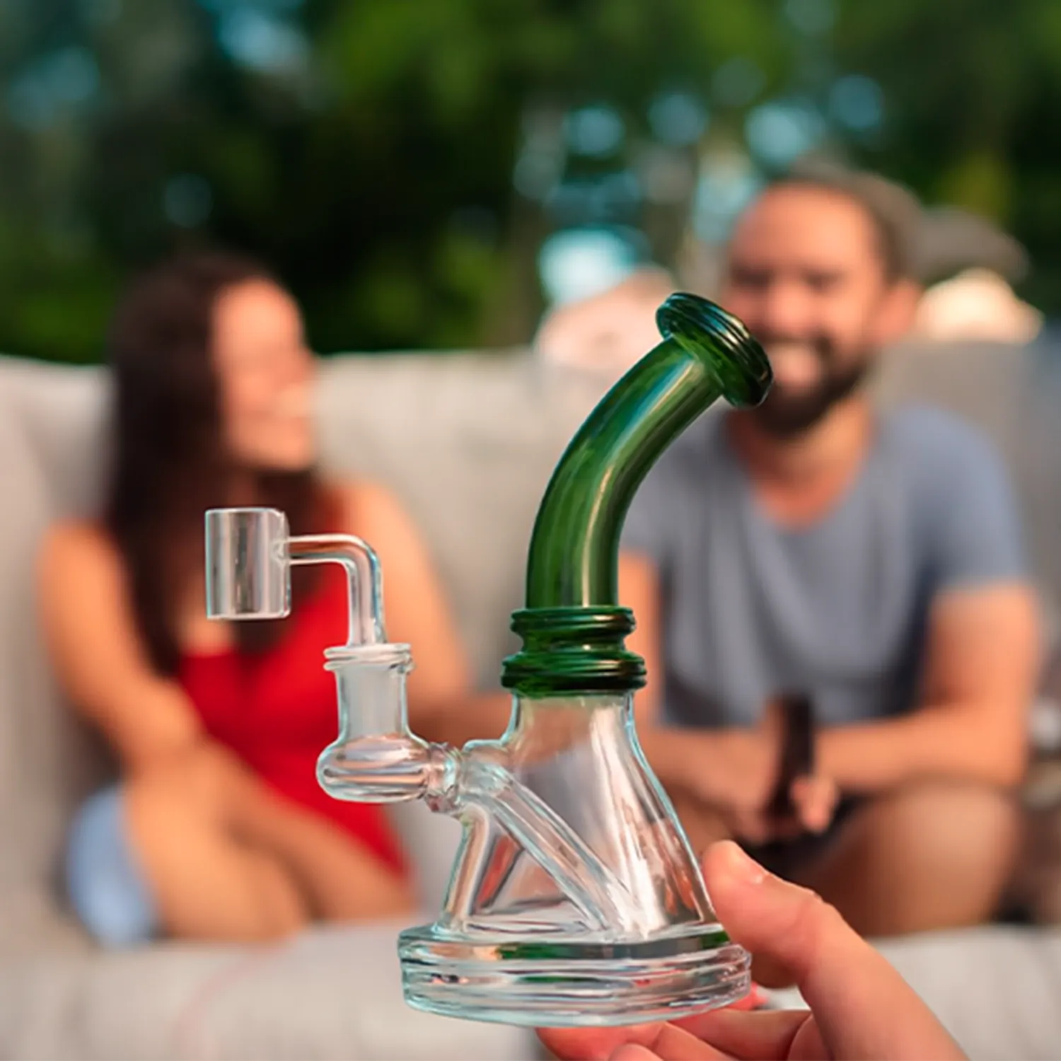 Close-up front view of a hand holding the 7" Mini Bent Neck Bong - Green showing its curved green neck and clear glass base against a blurred outdoor background with two people.