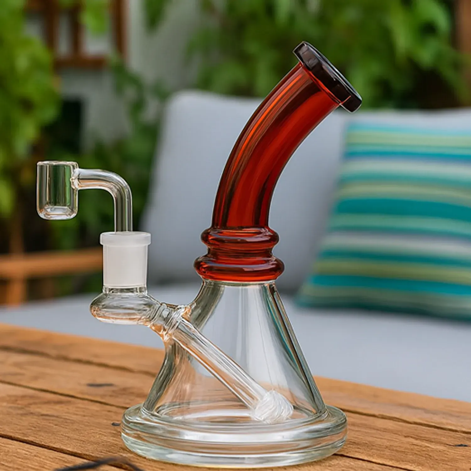 Three-quarter view of the 7" Mini Bent Neck Bong - Amber showing its amber bent neck and clear cone base on a wooden table with a blurred patio cushion background