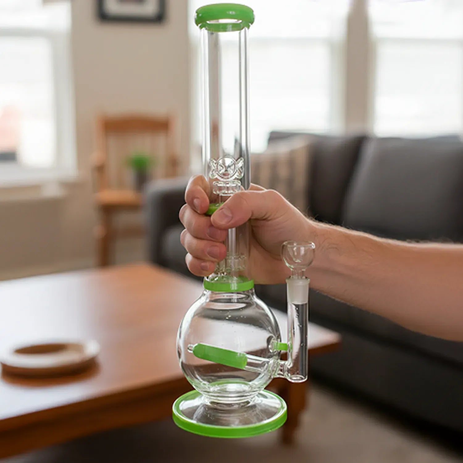 Handheld front view of the DHC Inline Glass Ball with UFO & Circ Perc Bong - 16 in., showing clear glass and green accents against a softly lit living room background