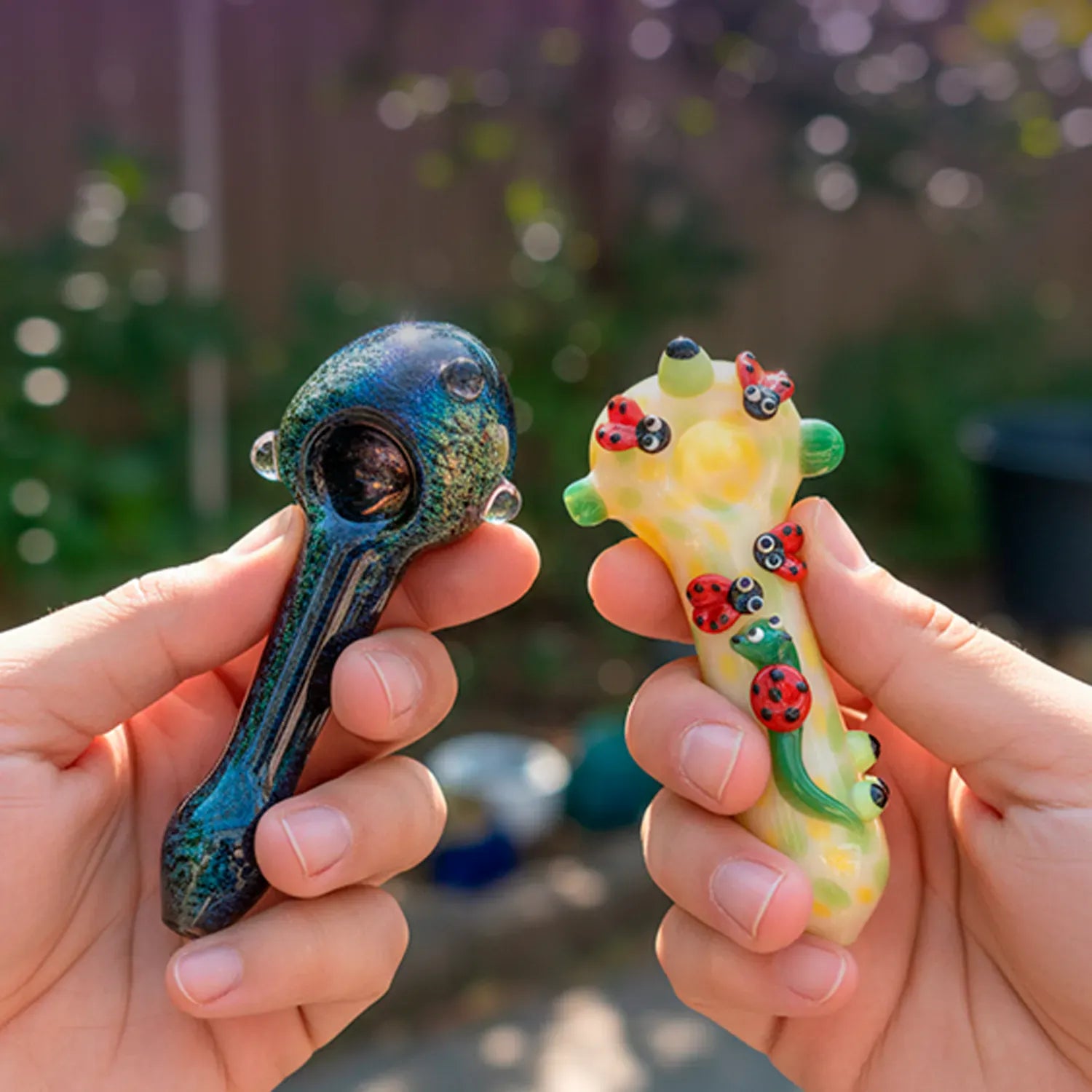 Close-up of two hand-held glass pipes, one a greenish-blue spoon and the other the DHC Fancy Ladybug and Snail Spoon Pipe - 6 in. decorated with tiny red ladybugs and a green snail.