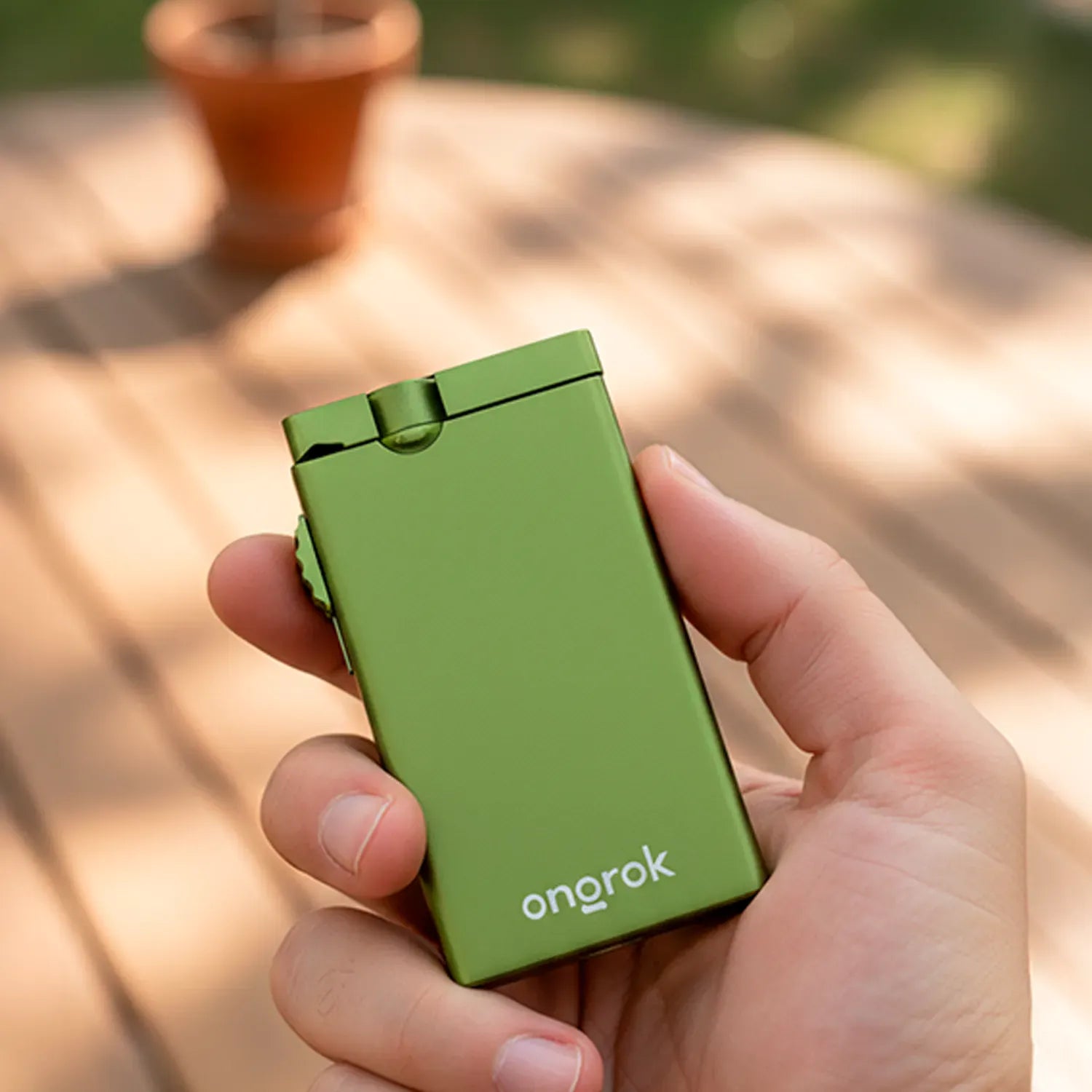Close-up of a hand holding a green Aluminum Dugout Pipe with a visible hinge and the "onorok" logo against a sunlit blurred wooden deck background