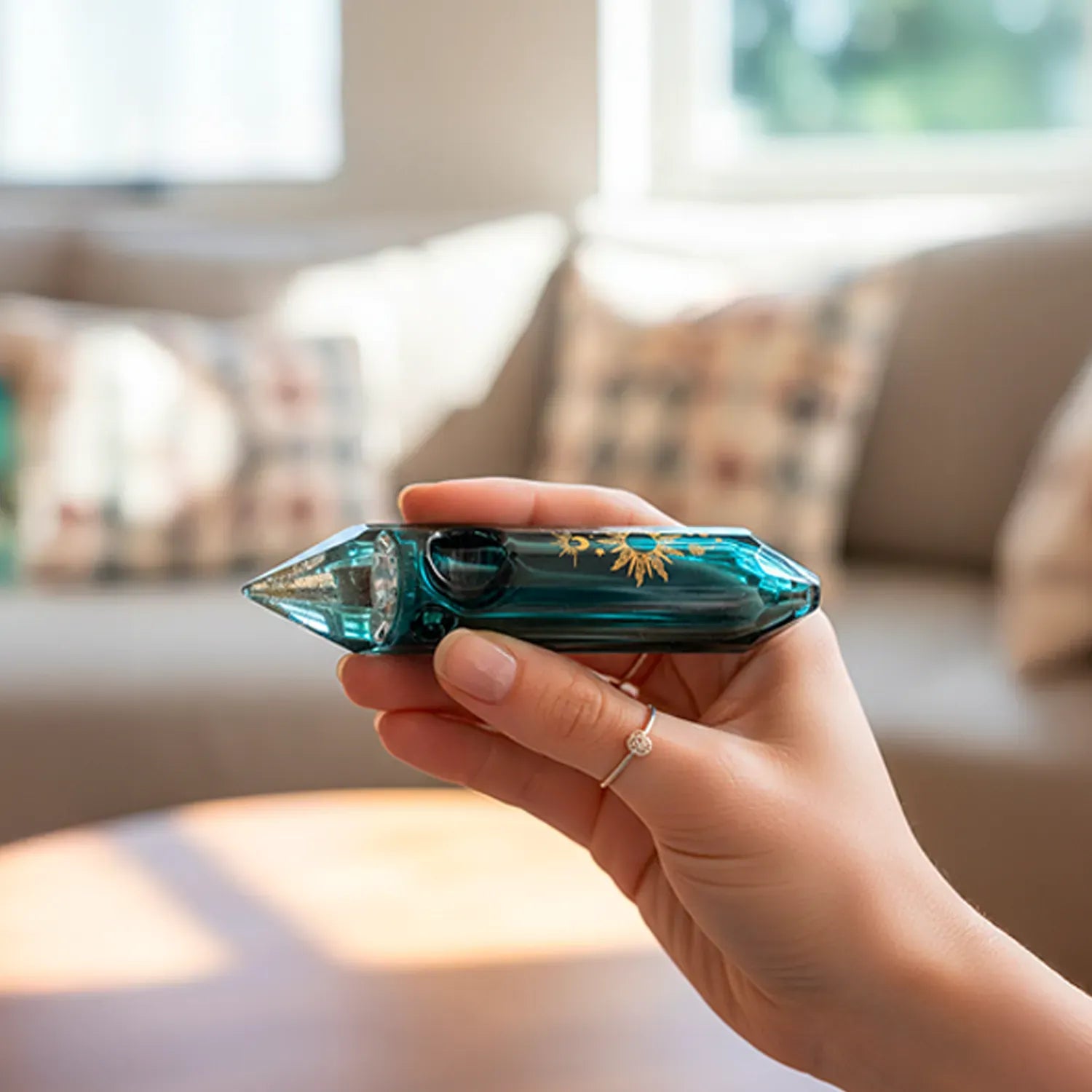Handheld side view of the Famous X Astral Power Crystal Hand Pipe - Black - 5" showcasing a teal-green faceted crystal shape with gold sun decals against a blurred living room background