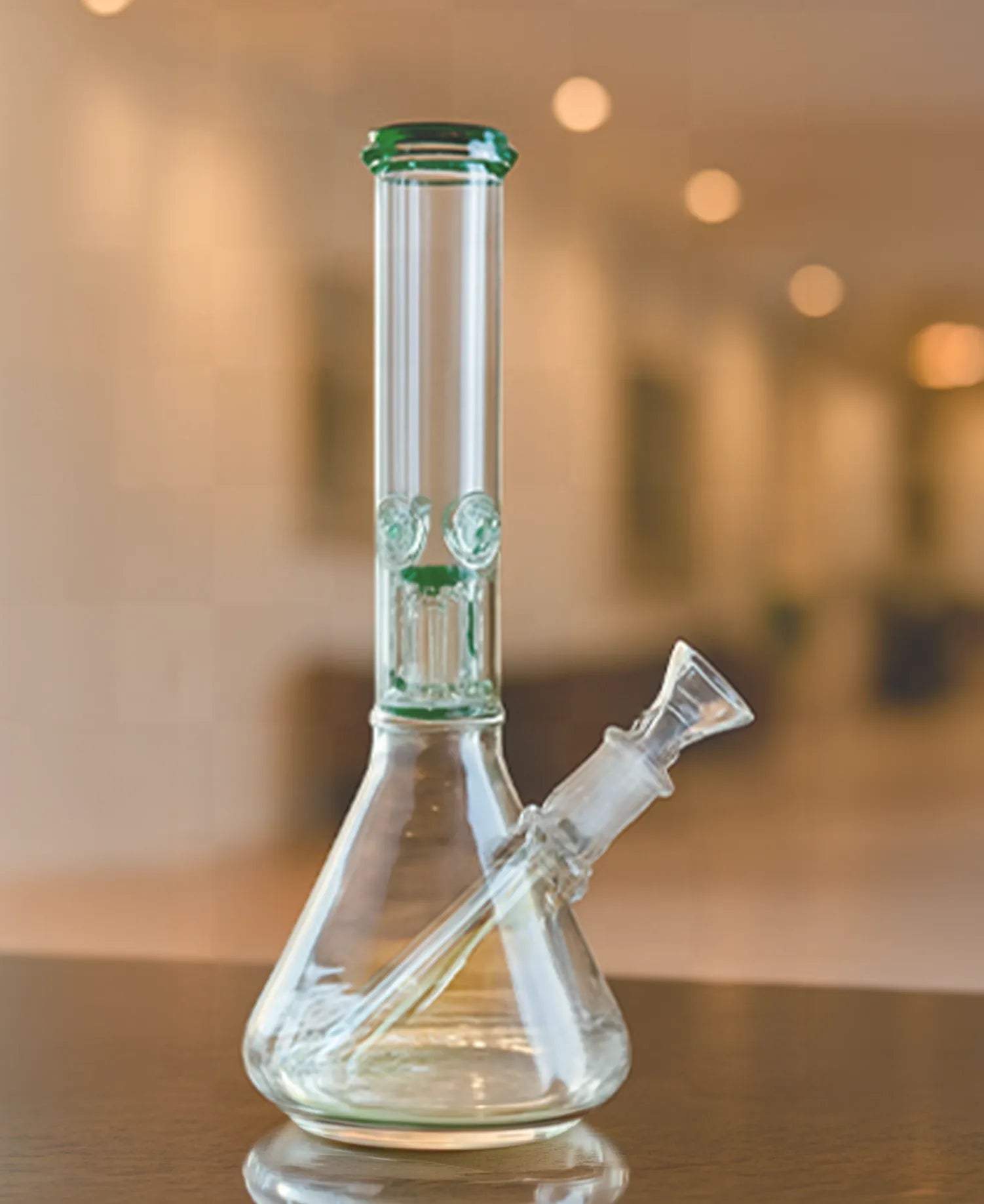 Angled close-up of the clear glass The Stash Shack 6 Arm Perc Beaker Bong on a wood table with warm bokeh lights behind it