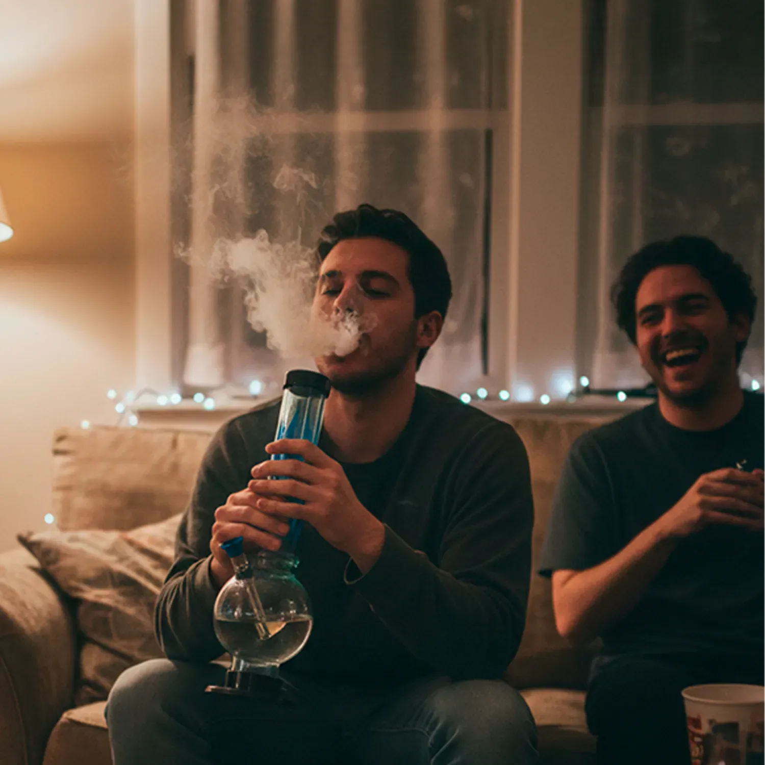 Center-frontal view of a man exhaling smoke while holding the JM Plastics Flame Bubble Base Acrylic Bong - 12 in., the red-accented tube and round water base lit by warm room lighting.
