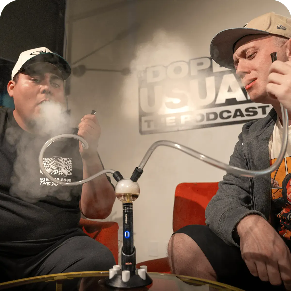 Two men seated on red chairs exhale clouds while using the Cipher NOVA Water Hookah Kit on a glass table between them.