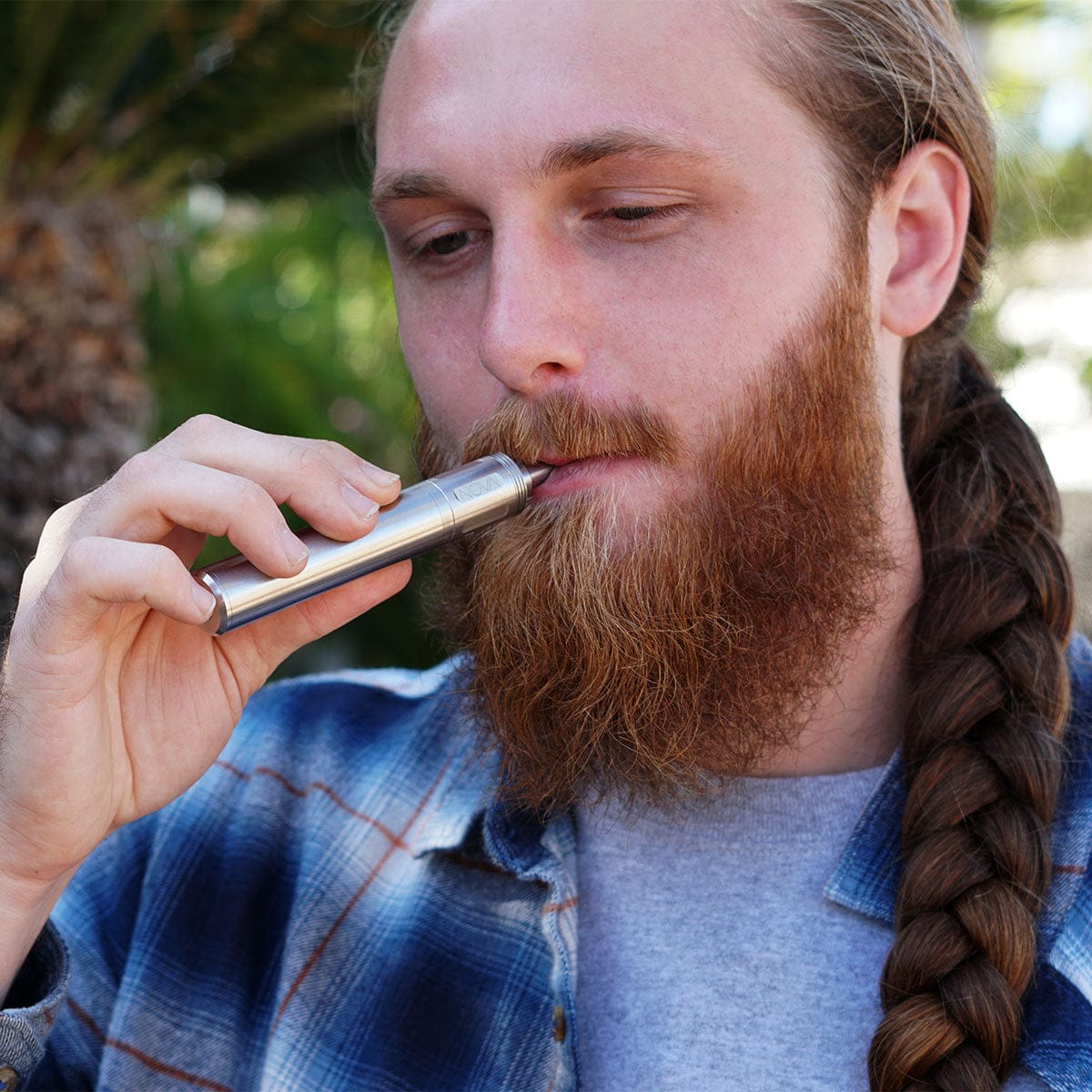 Close-up of a bearded man holding the Cipher NOVA | Iridescent - V2 vaporizer to his lips, shot from a slight side angle with a blurred outdoor background.