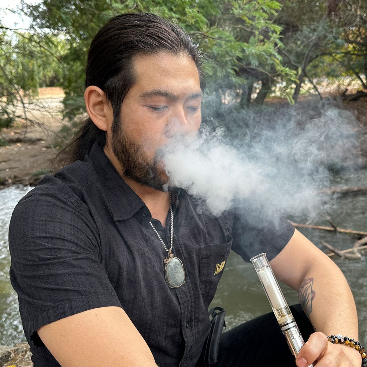 Three-quarter view of a seated man exhaling a thick cloud while holding the Cipher NOVA | Iridescent - V2 vape near a tree-lined creek.