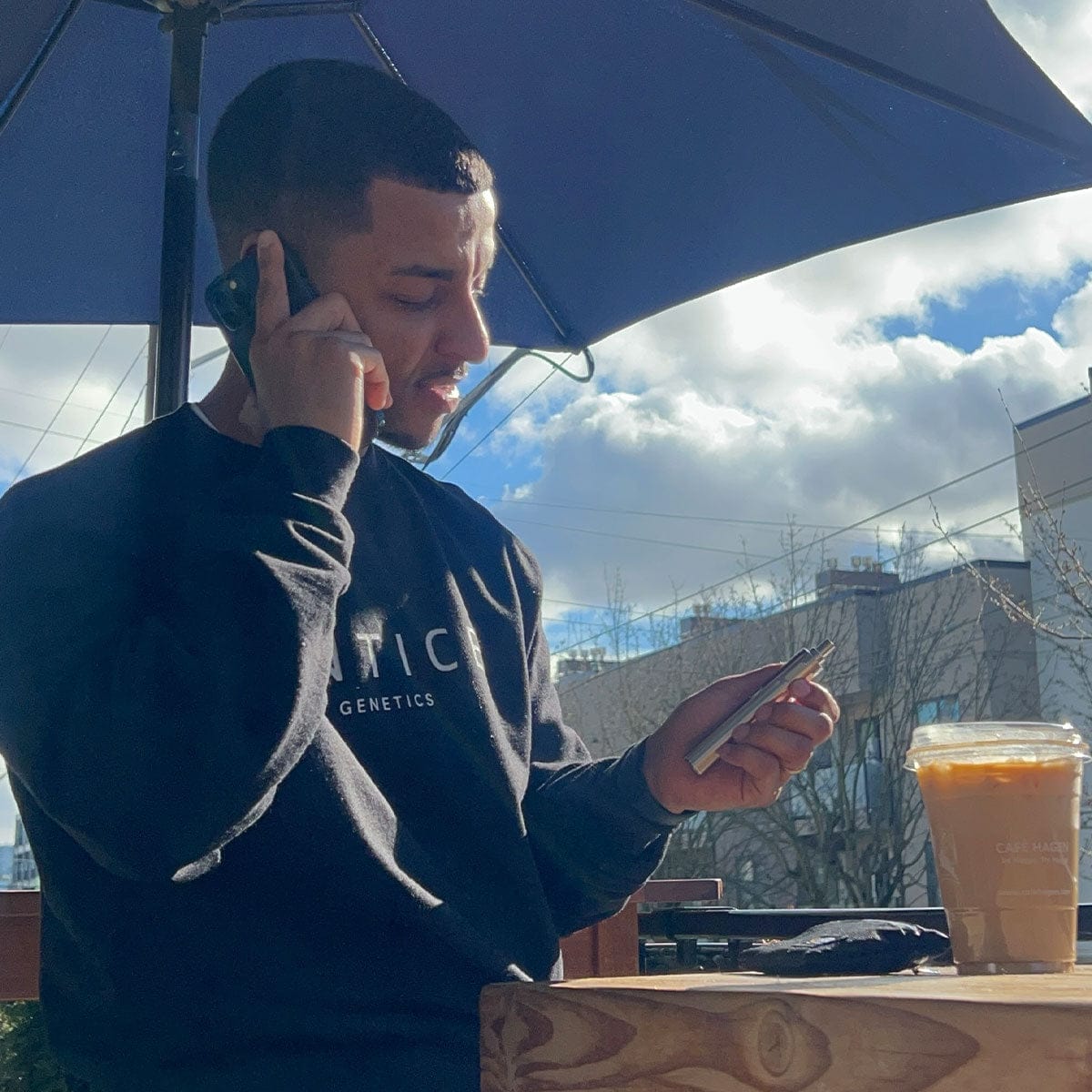 Side view of a person under a blue patio umbrella holding a silver Cipher NOVA | Carbon Black - V2 device while looking at it, with a cold coffee on the table and sunlit clouds in the background