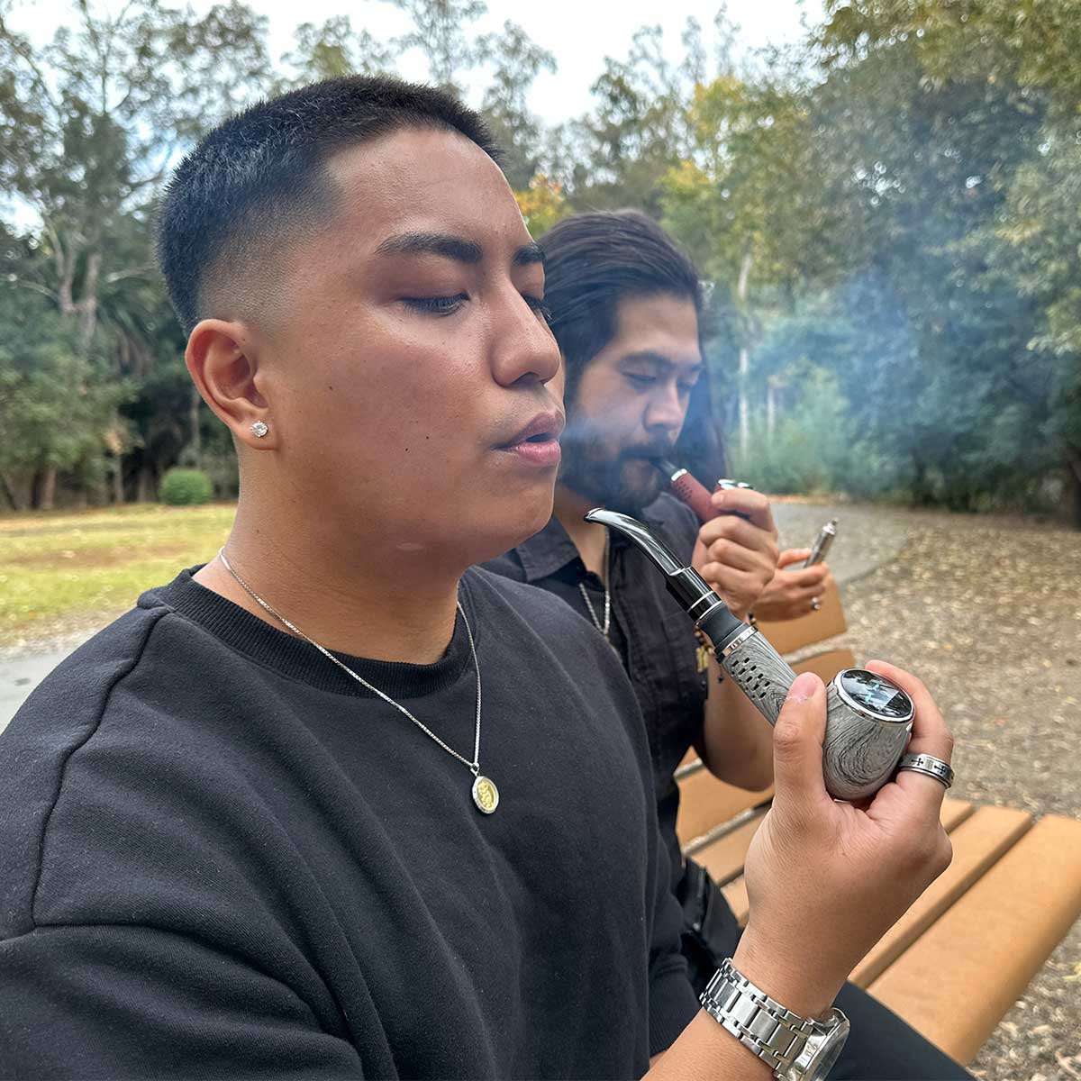 Close-up side view of a person holding and inhaling from the Cipher Nautilus V3 Churchwarden Stem Kit in a gray woodgrain finish while seated on a park bench with trees and another person smoking in the background
