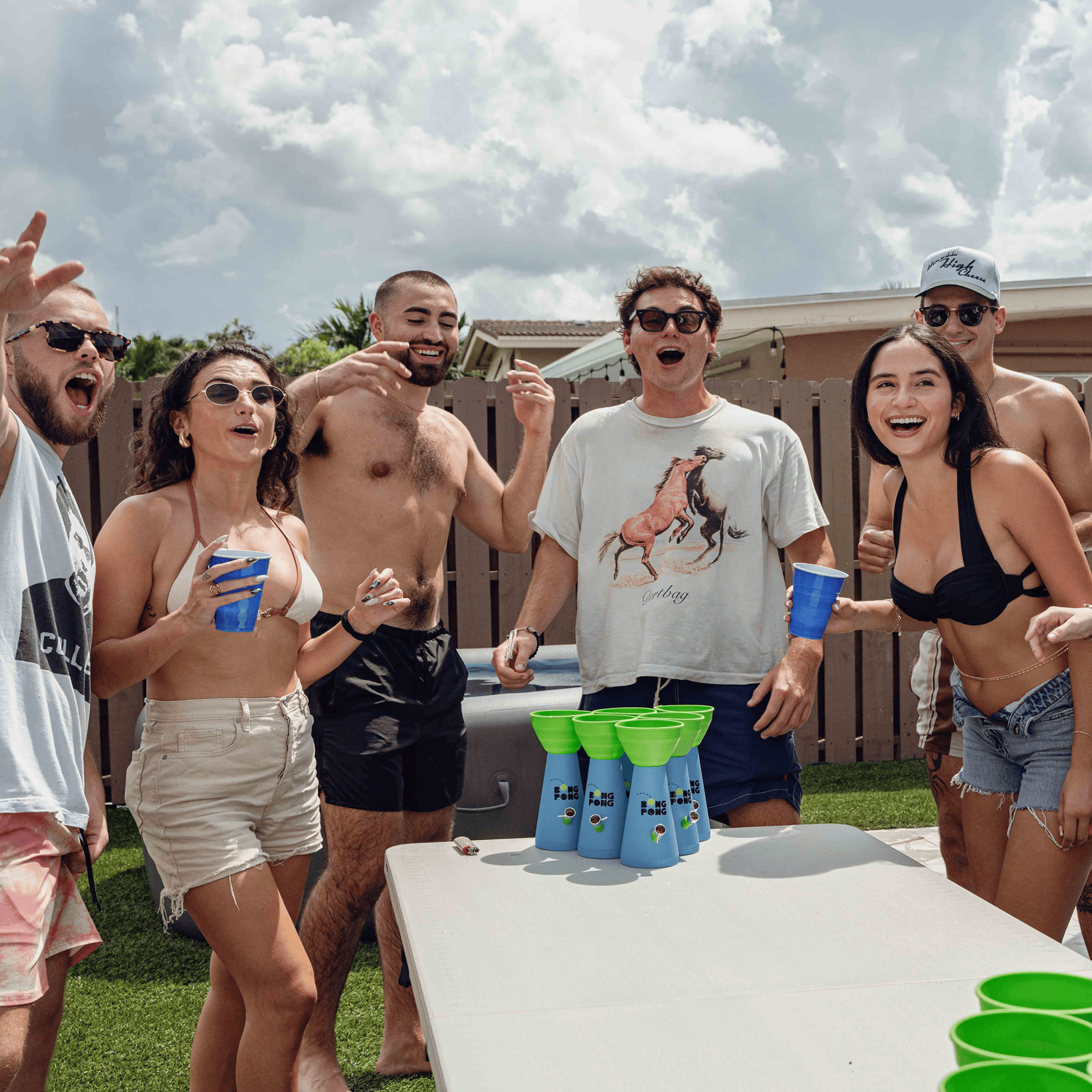 Group shot from a low angle showing friends cheering around a backyard table with a stack of blue-and-green Bong Pong OG funnels in the foreground