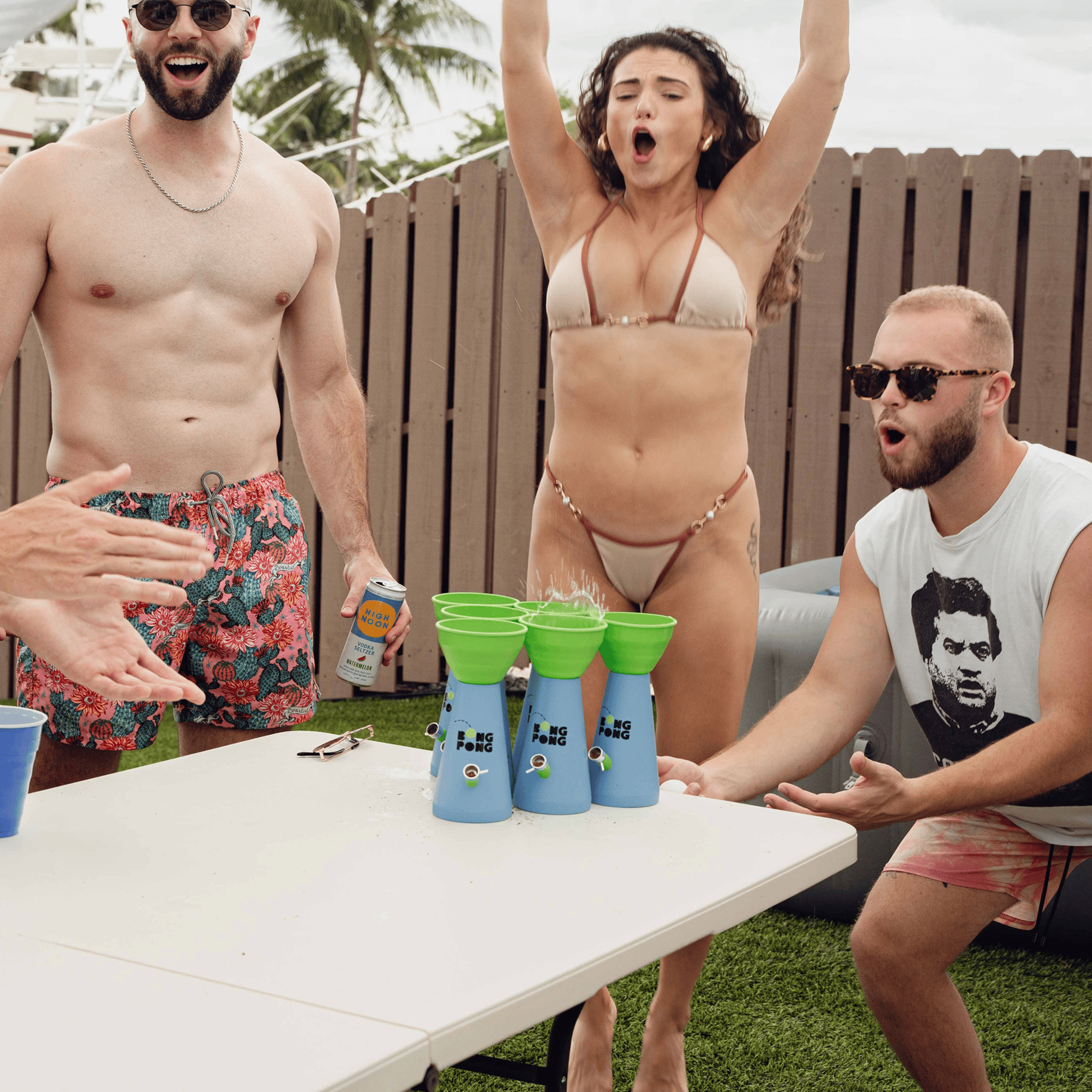 Three-quarter view of a blue and green stacked game tower in the foreground with friends cheering behind it, labeled Bong Pong OG