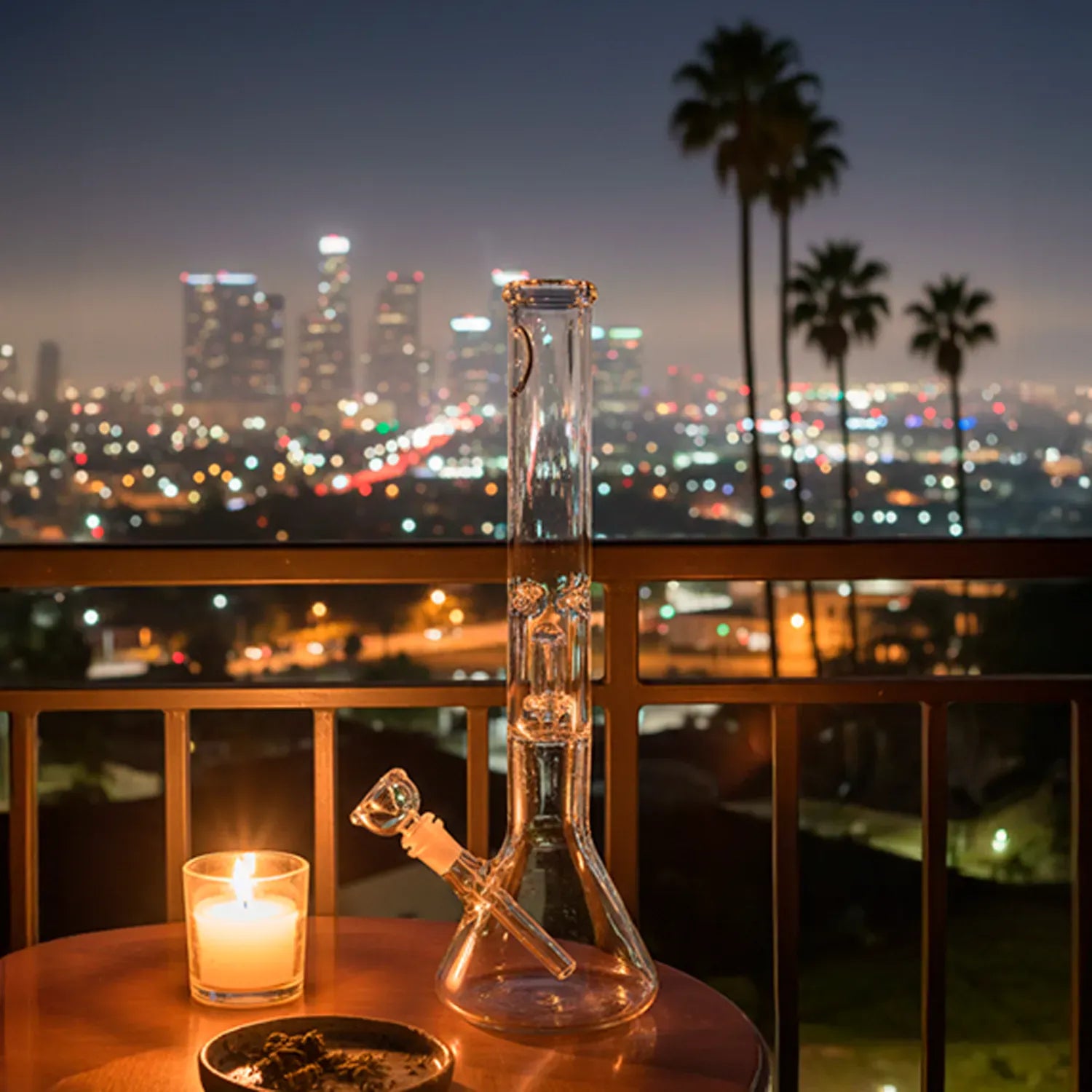 Centered front view of the "Magnum Flow" Thick Glass Beaker Showerhead Perc Bong on a balcony table lit by a candle with a blurred city skyline and palm trees at night
