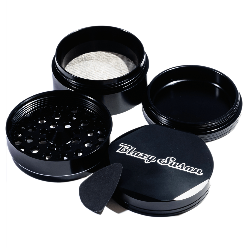 Angled top-down view of the opened Blazy Susan 4-pc 3 Chamber Grinder showing its stacked black metal chambers, perforated grinding plate, mesh screen, lid with logo, and small black scraper.