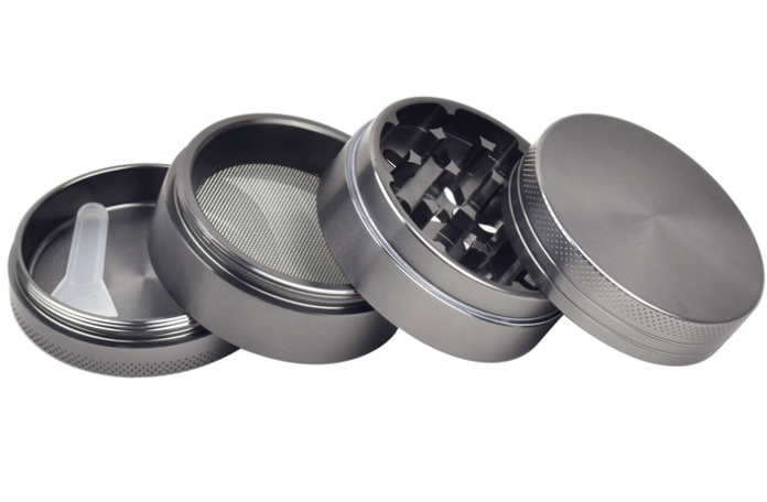 Angled close-up of a Gunmetal 4-Piece Aluminum Grinder showing its four separated chambers, mesh screen, sharp diamond teeth, and small plastic scraper on a white background