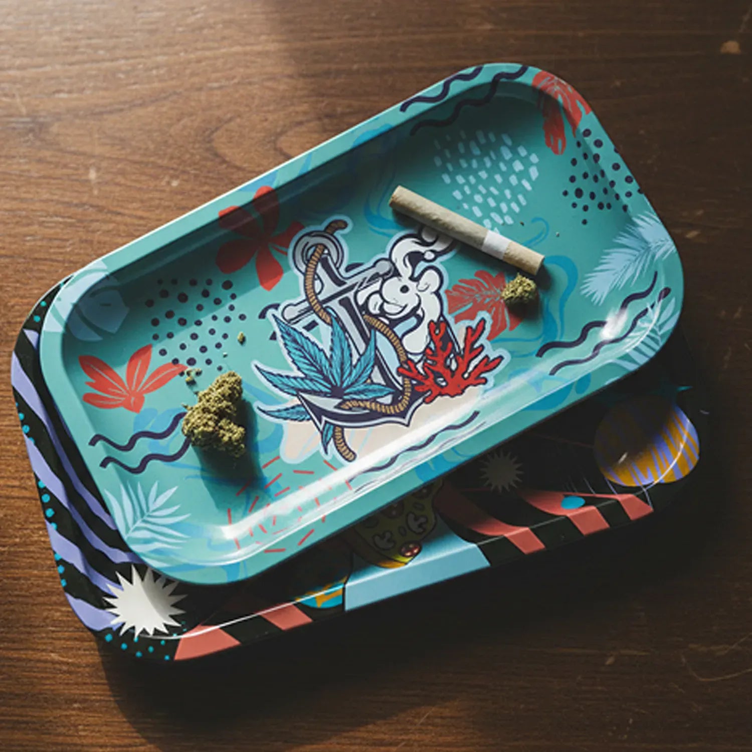 Top-down view of the Atomik Tray - Medium Explorer showing a teal tray with anchor and leaf artwork, a rolled joint and small herb pieces on a wooden table.