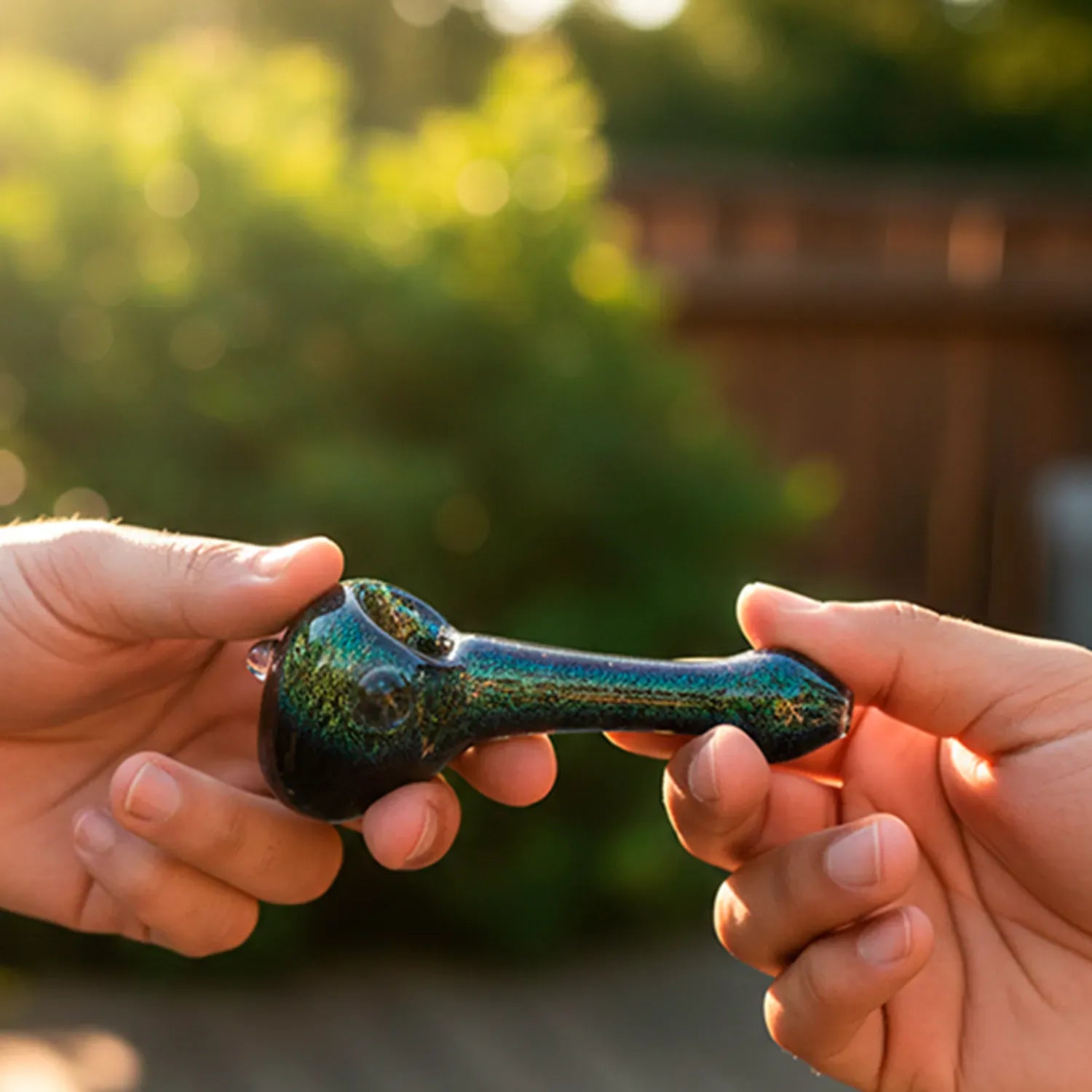 Hands passing a shimmering blue-green Galactic Storm Full Dichro Spoon Hand Pipe in a sunlit outdoor setting, close-up on the bowl and stem.