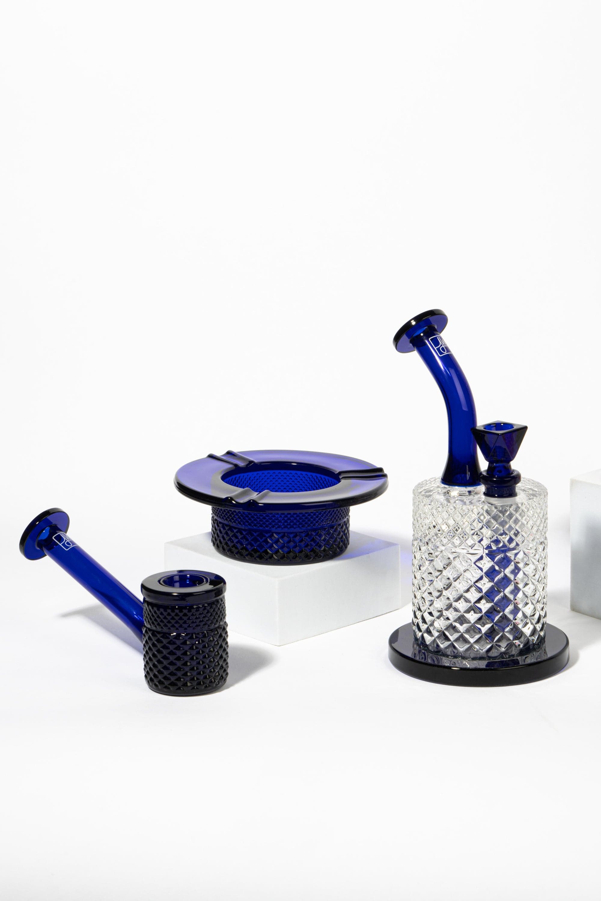 Close-up of the cobalt Twenties Collection Bong | Cobalt set photographed on white blocks, showing a tilted neck, textured clear glass base, and matching blue accessories