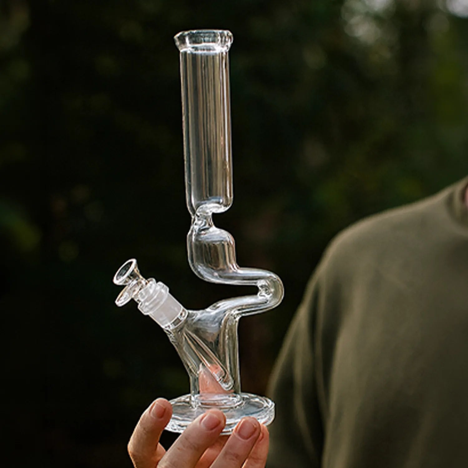 Close-up of a hand-held DHC Fumed Zong Bong - 15 in. showing the clear, curving S-shaped neck and straight cylindrical mouthpiece against a blurred dark-green background