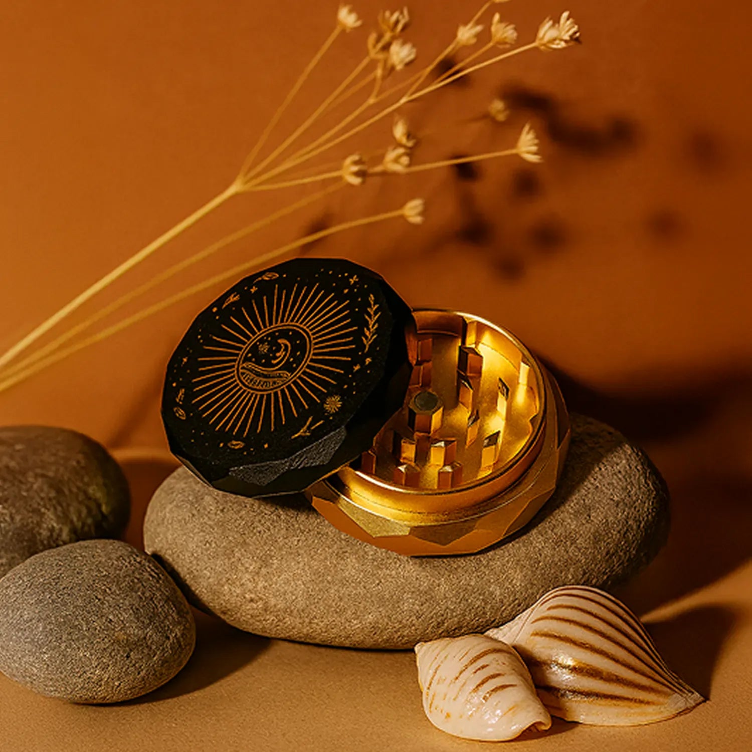 Close-up three-quarter view of the Famous X Astral Power Aluminum 2 Stage Grinder -Black and Gold resting open on smooth stones, its black lid with gold sunburst engraving and shiny gold interior lit warmly.