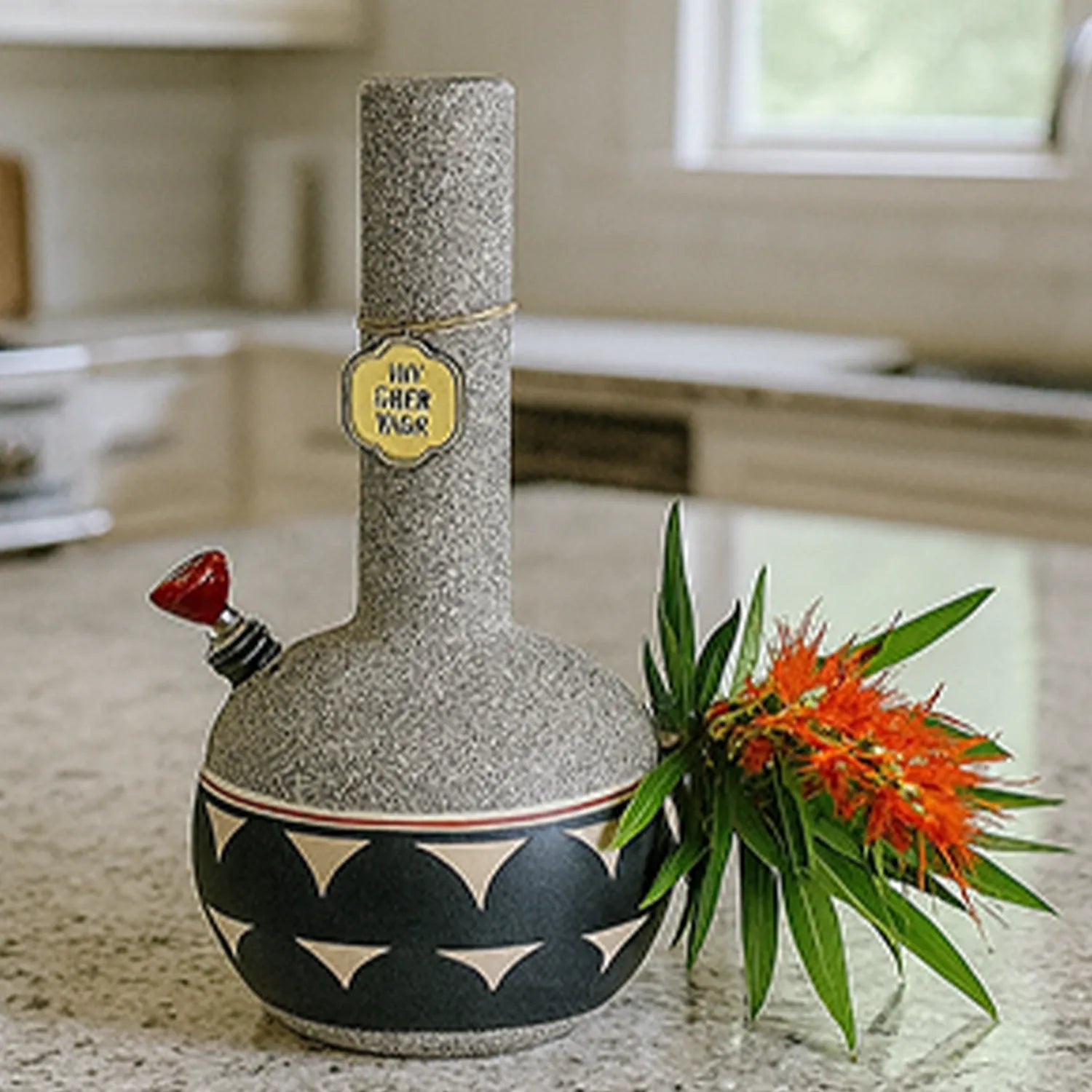 Front three-quarter view of the speckled, round-bodied My Bud Vase "Coyote" Bong with geometric banding and a red-tipped downstem, staged on a kitchen counter beside orange flowers.