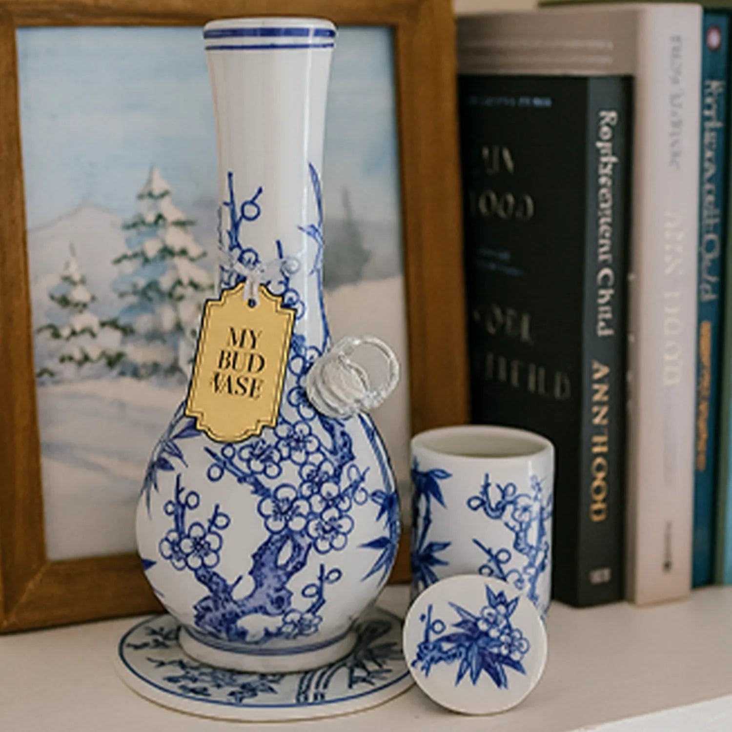 Close-up of the My Bud Vase "Luck" Chinese Porcelain Vase Bong showing its blue floral pattern on white ceramic with a gold tag, small cup, and books in soft light