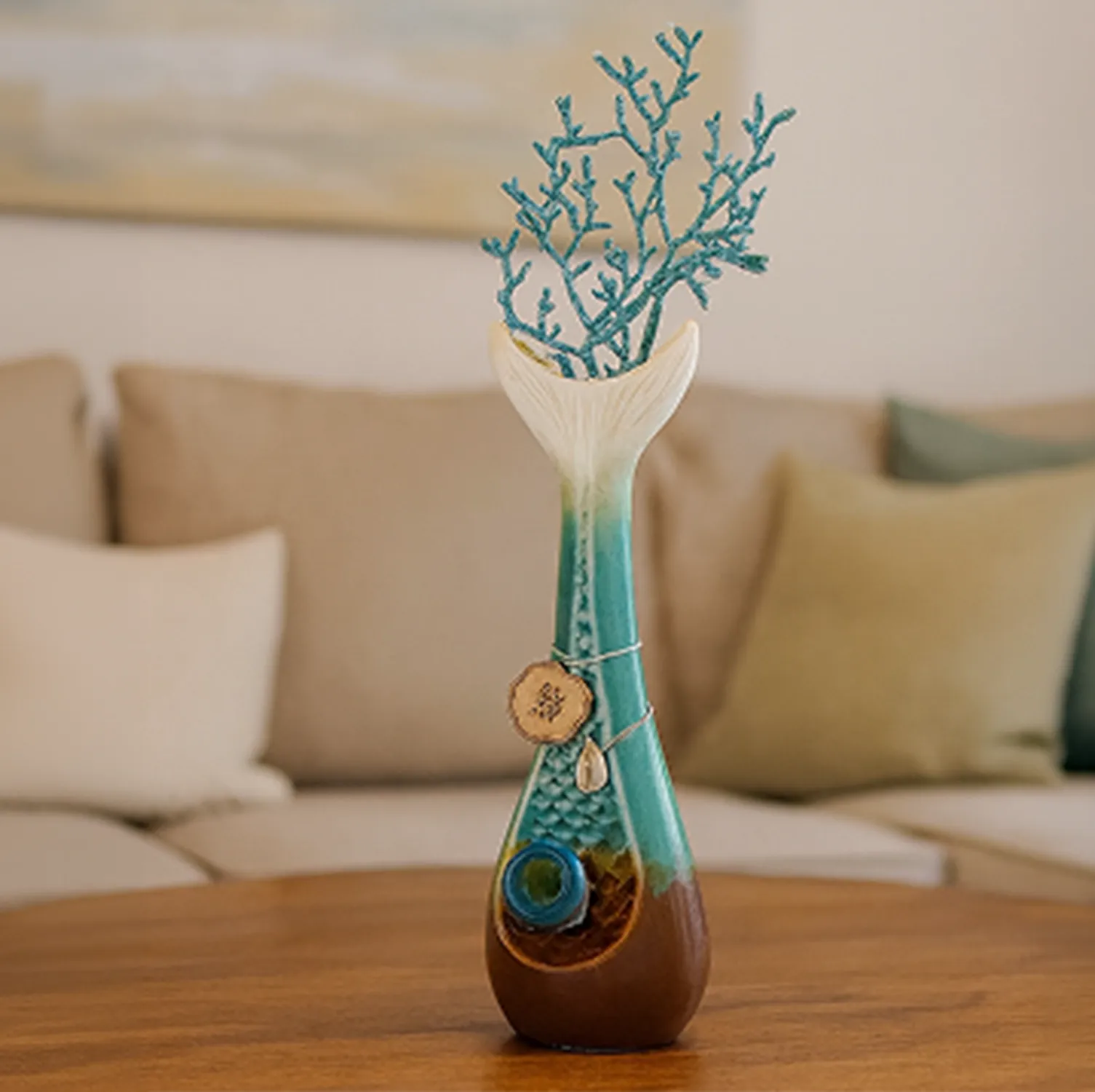 Close-up front view of the My Bud Vase "Mermaid" Bong showing its teal mermaid-tail neck and decorative coral sprig against a blurred beige sofa background.