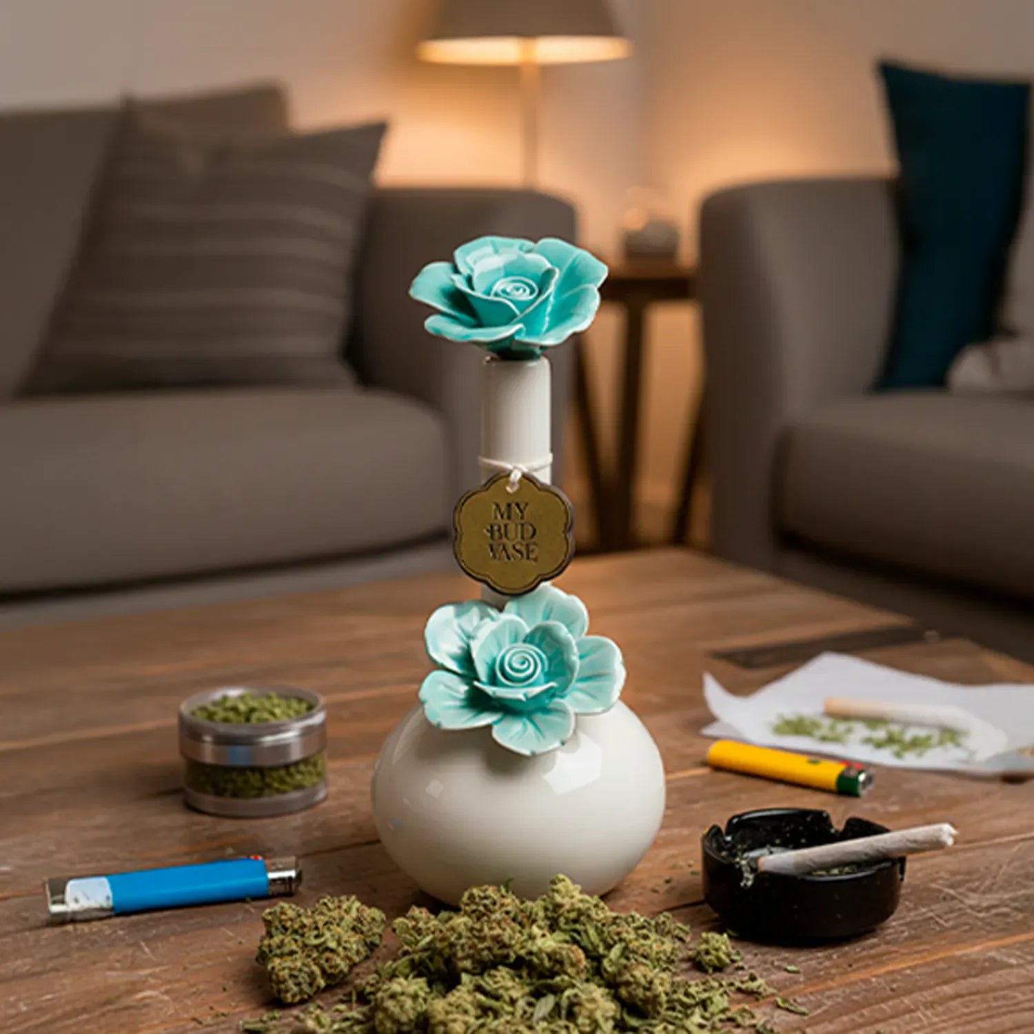 Centered front view of the My Bud Vase Bong - Rosette showing a white round vase with two mauve-green rosette flowers on a wooden coffee table in warm lamp light