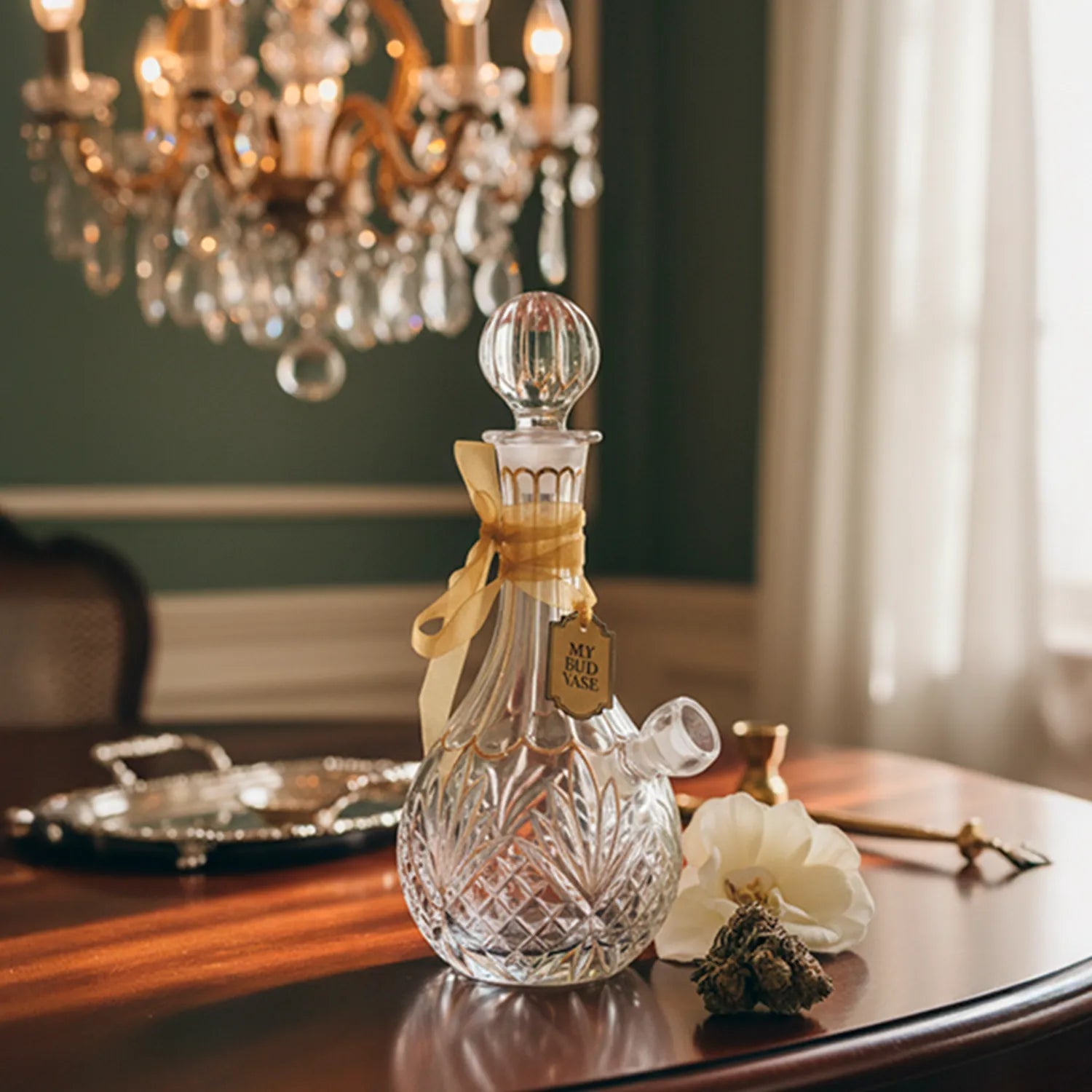 Close-up of the glass decanter-style My Bud Vase "Stardust" Bong on a polished wooden table, ribbon-tied neck and chandelier bokeh background.