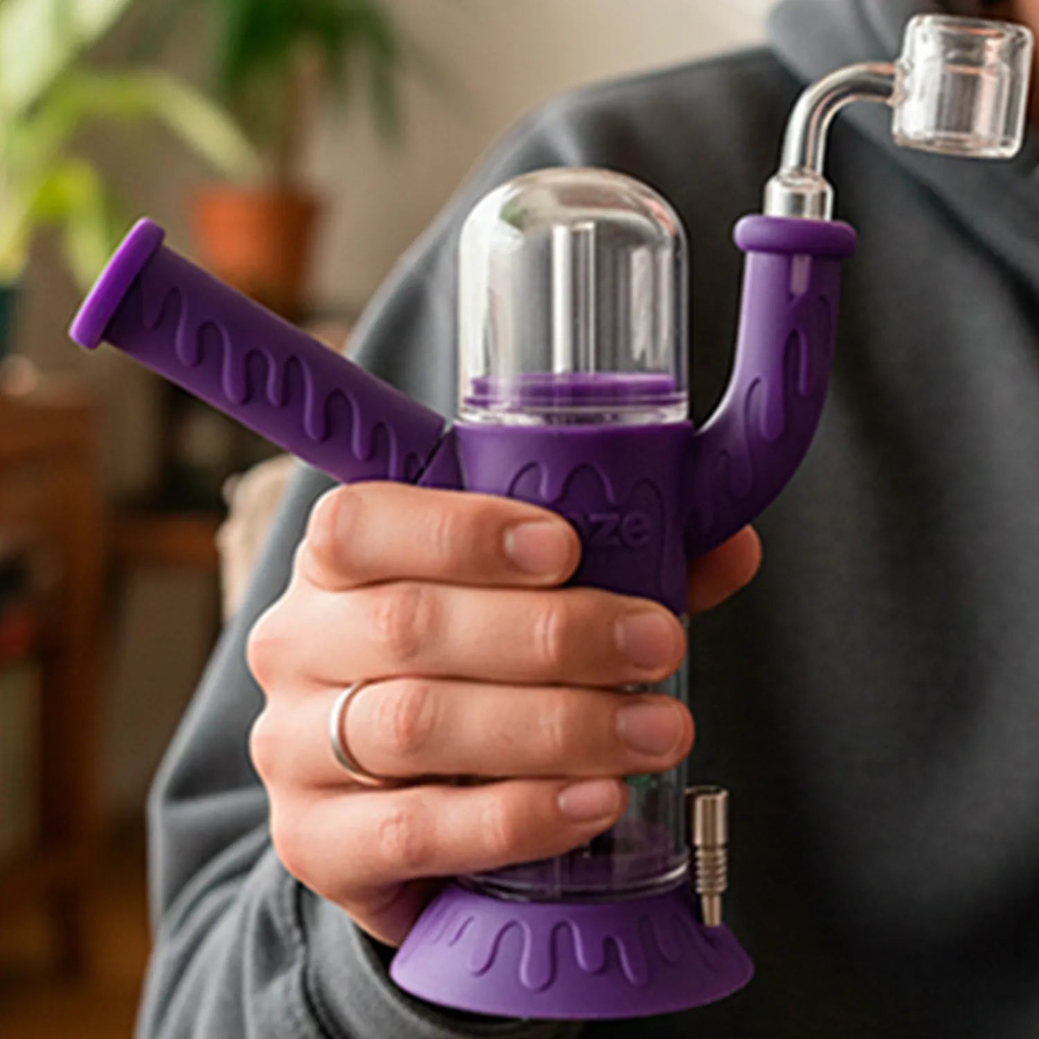 Close-up of a hand holding the Ooze Cranium Silicone 4-in-1 Hybrid Bong from a front angle, showing its purple silicone dripped design, clear dome chamber, and glass vapor joint.