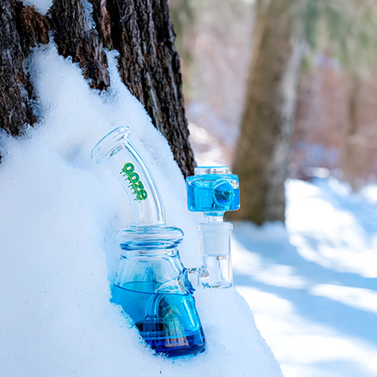 Close-up of the Ooze Glyco Bong Glycerin Chilled Glass Bong half-buried in snow against a tree trunk, blue glycerin visible.