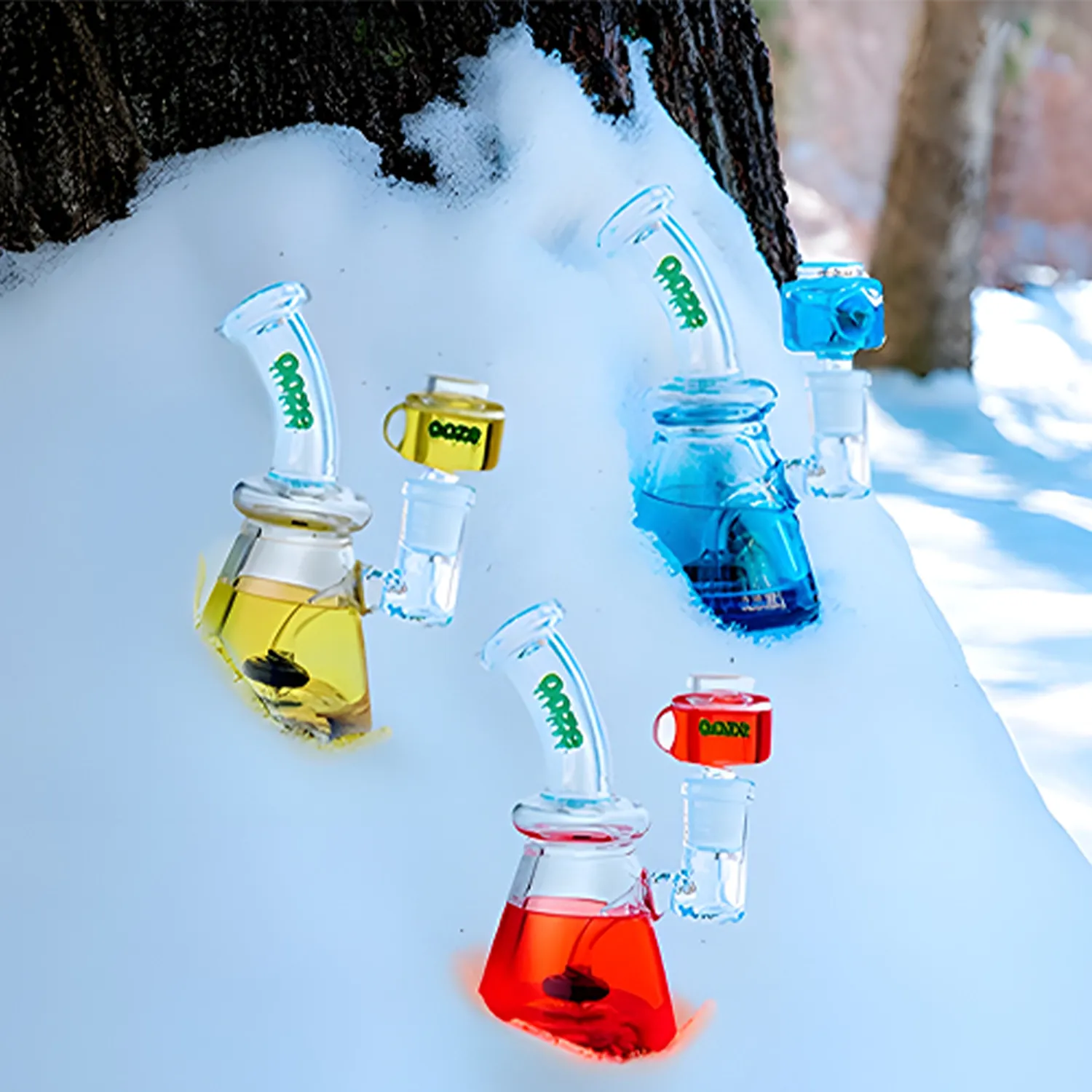 Diagonal close-up of three colorful Ooze Glyco Bong Glycerin Chilled Glass Bong units (yellow, blue, red) embedded in snow against a tree backdrop.