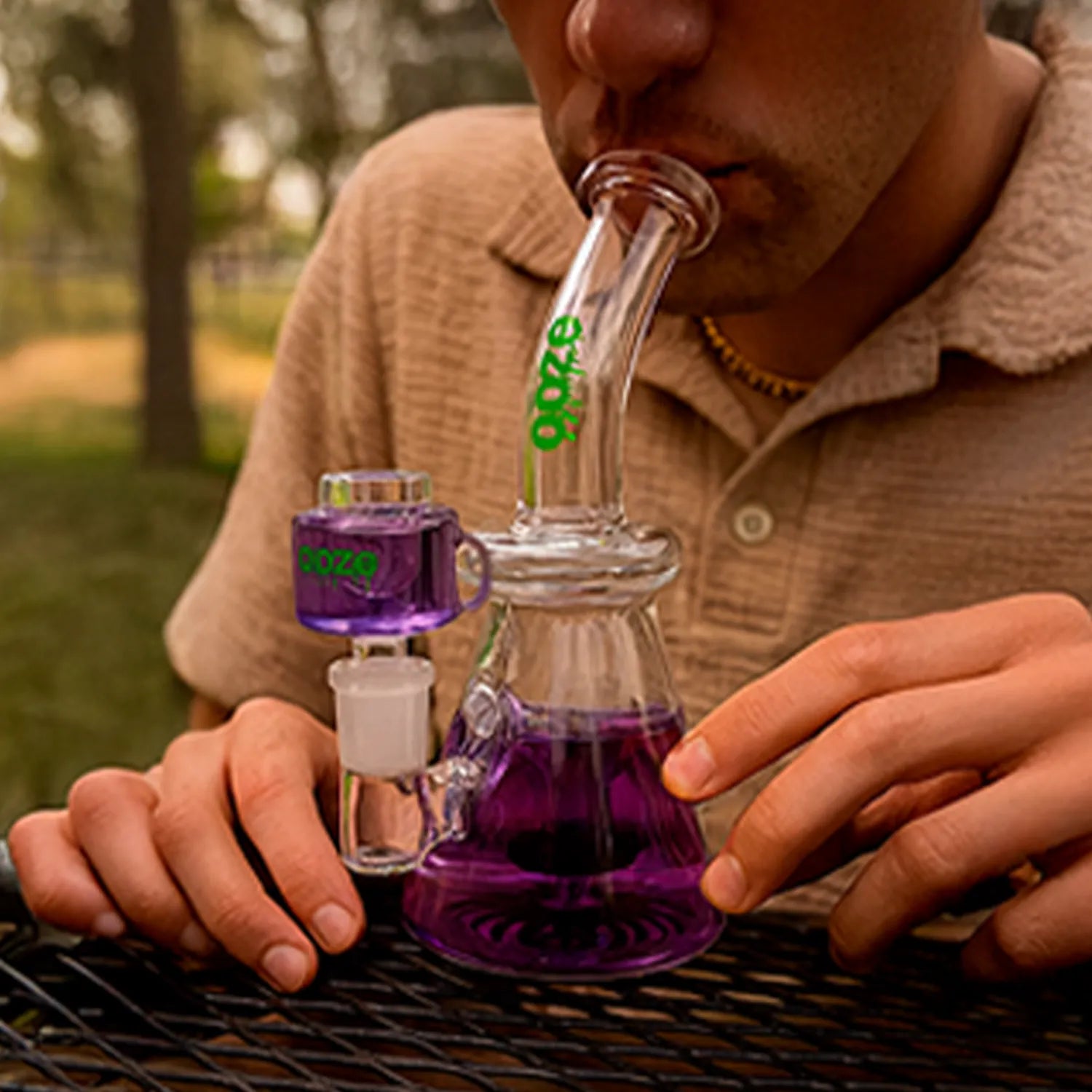 Close-up of a person using the Ooze Glyco Bong Glycerin Chilled Glass Bong, shown at mouth-to-neck angle with purple water and outdoor background