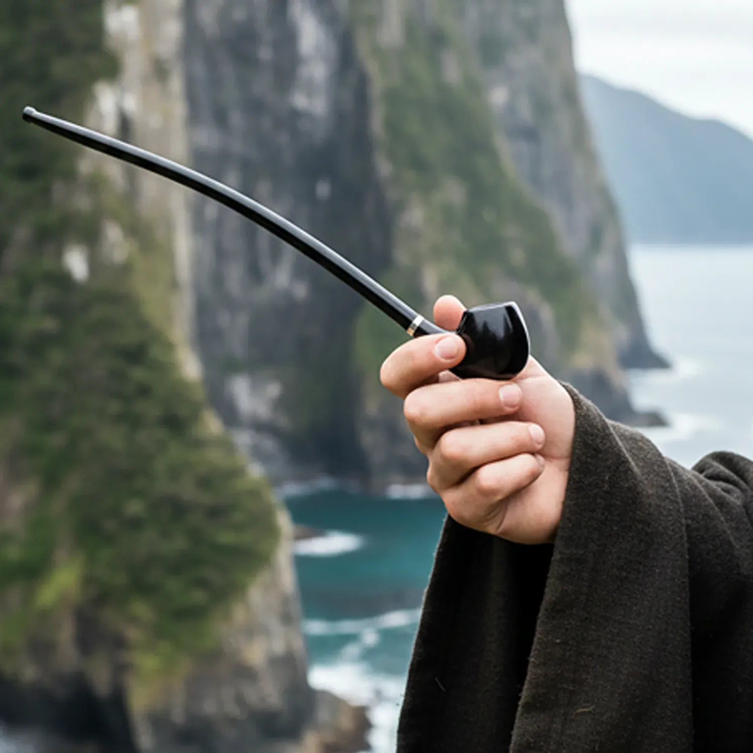 A close-up of a hand holding the Pulsar Shire Pipes Deep Churchwarden Tobacco Pipe with its long black stem arcing left against a misty coastal cliff backdrop