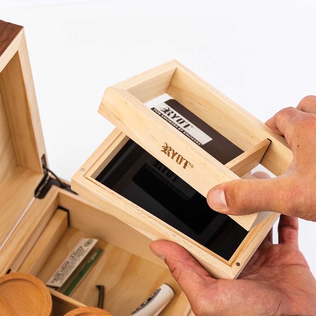 Top-down view of hands lifting the small inner tray of the Ryot Humidor Walnut Combo Box, showing the light wood grain, black glass humidifier insert, and open compartment below.