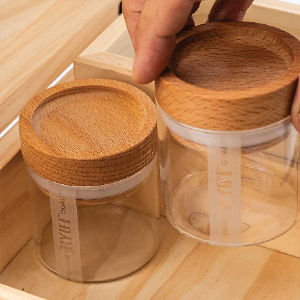 Close-up, top-down view of two clear jars with walnut lids inside a wooden Ryot Humidor Walnut Combo Box, one lid being lifted by fingers.