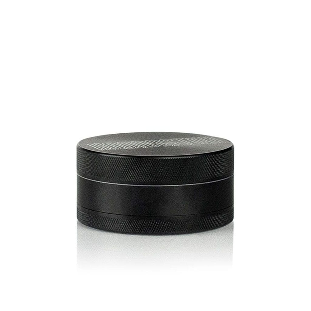 Low-angle front view of the black Ryot 4Pc Jar Body Grinders sitting on a reflective white surface, showing its textured lid and layered metal body.