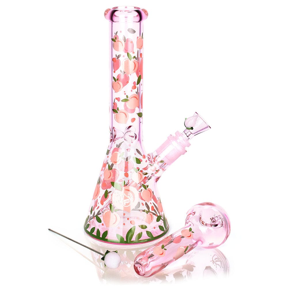 Front view of the pink Pulsar Fruit Series Peaches & Cream Herb Pipe Glow Duo - 10" / 14Mm F standing on a white background with peach motif and a matching handpipe and dab tool beside it