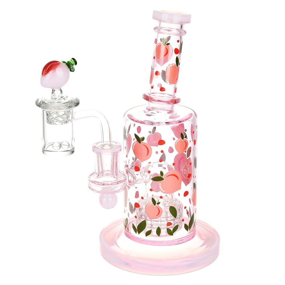 Angled front view of the Pulsar Fruit Series Peaches & Cream Glow In The Dark Dab Rig Kit - 8" / 14Mm F, a pink glass rig decorated with scattered peach motifs and a matching peach-topped banger against a white background.