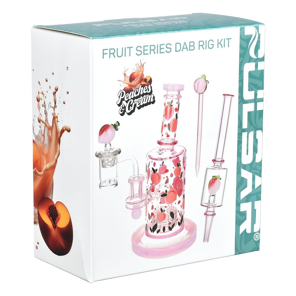 Angled front view of the boxed Pulsar Fruit Series Peaches & Cream Glow In The Dark Dab Rig Kit - 8" / 14Mm F showing the pink peach-patterned glass rig and accessories on white packaging.
