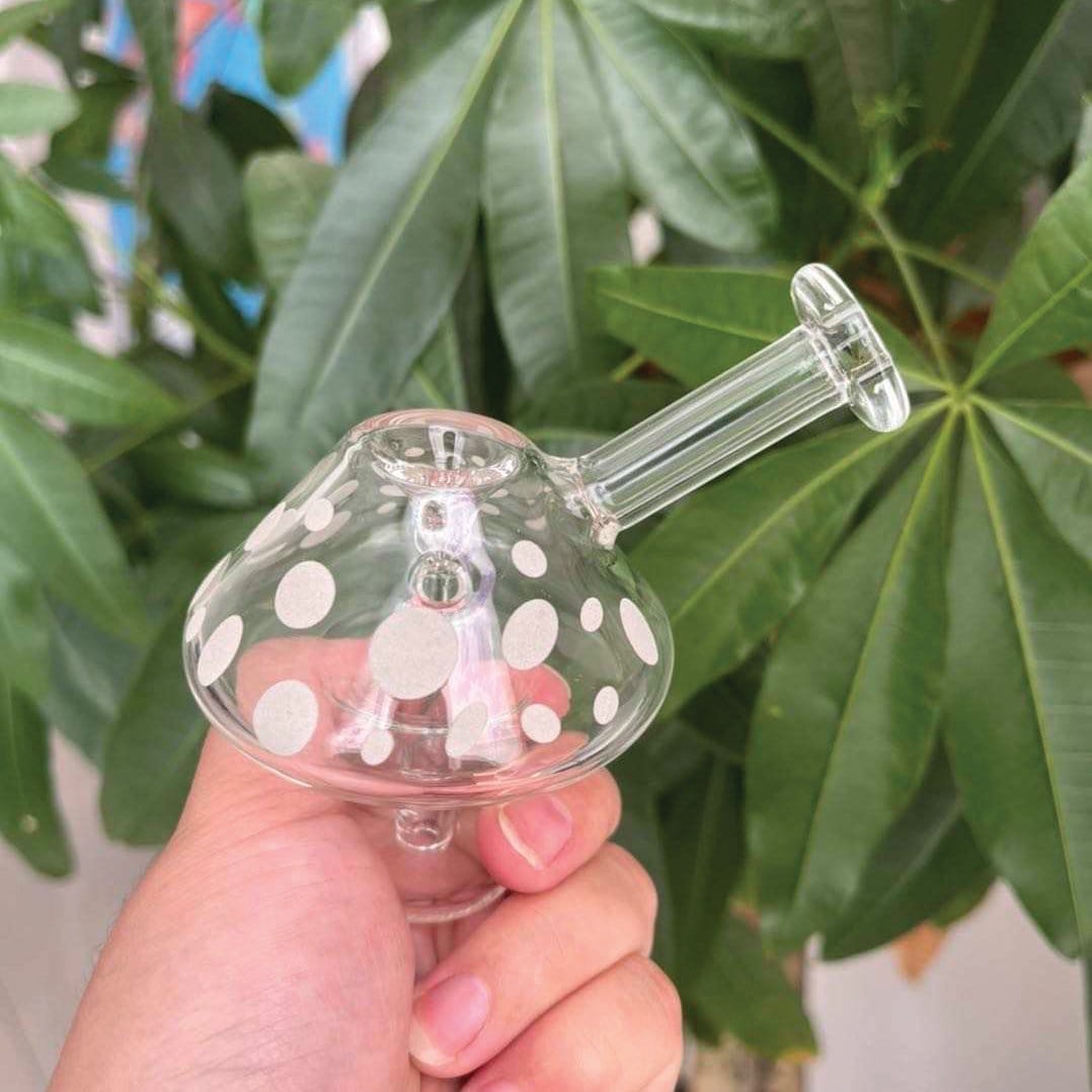 A hand holds the “Mushroom” Glow-in-the-Dark Mini Bubbler, showcasing its rounded shape against a blurred green leaf background.